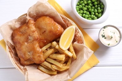 Tasty fish, chips, sauce, peas and lemon in wicker bowl on white wooden table, top view