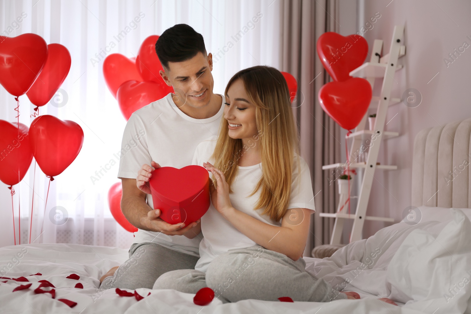 Photo of Man presenting gift to his girlfriend in room decorated with heart shaped balloons. Valentine's day celebration