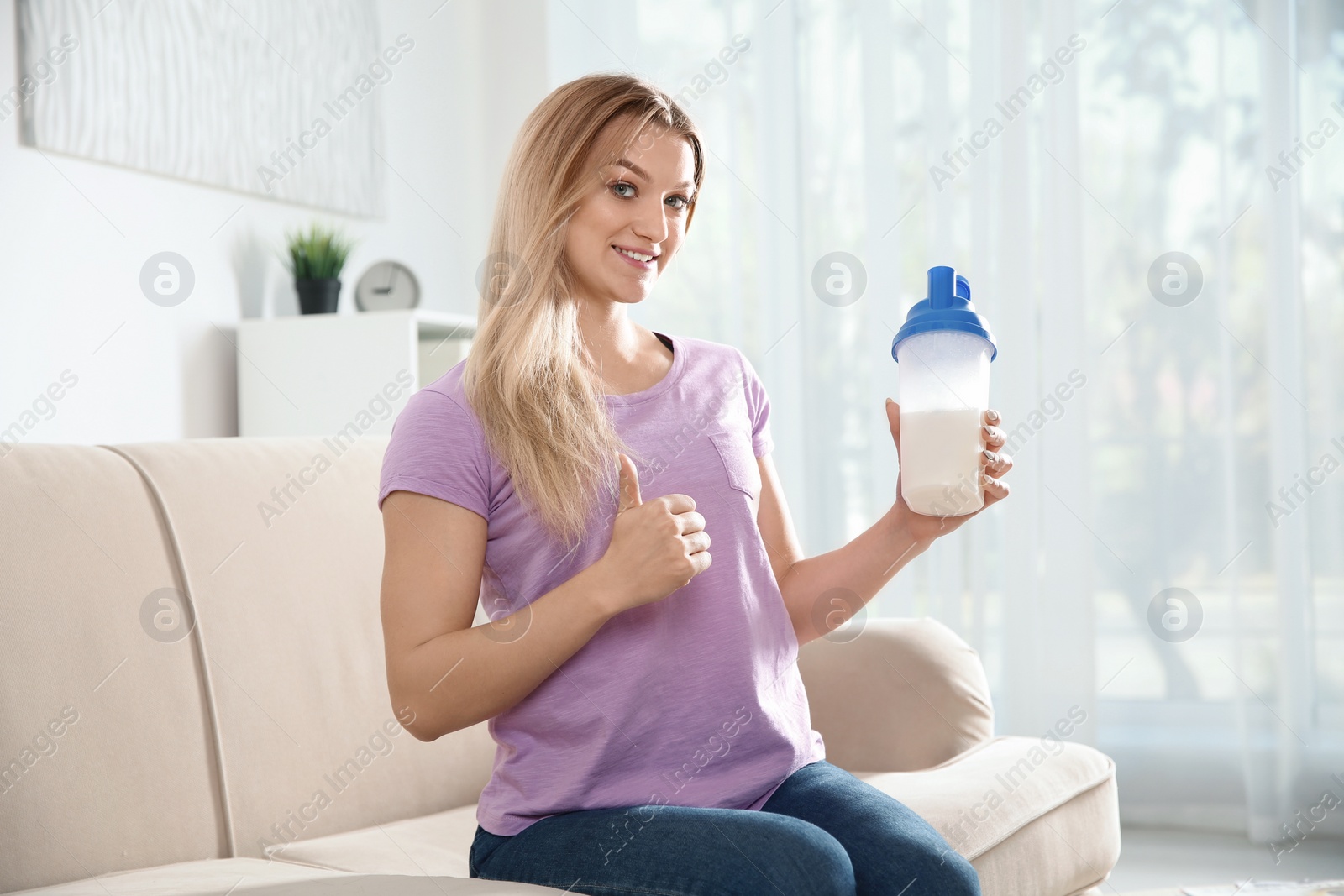 Photo of Young woman with bottle of protein shake sitting on sofa at home