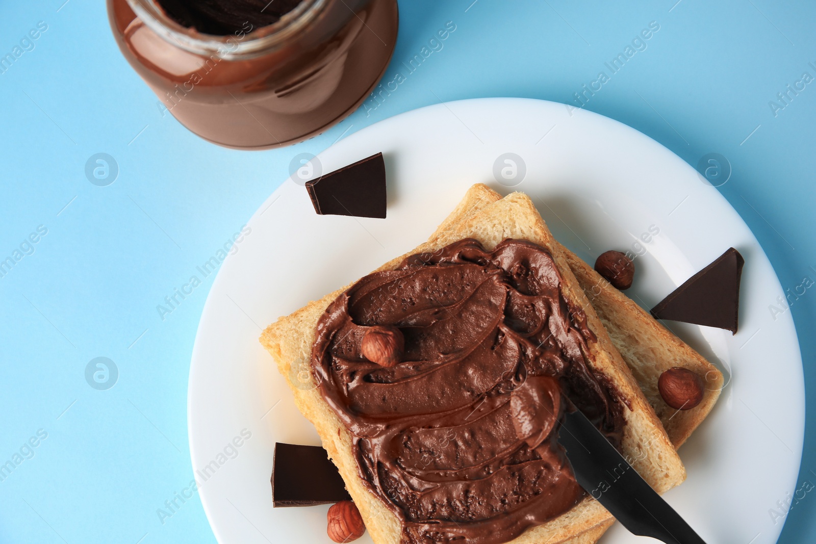 Photo of Spreading chocolate paste onto bread near jar of cream on light blue background, flat lay