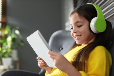 Photo of Cute little girl with headphones and tablet listening to audiobook at home