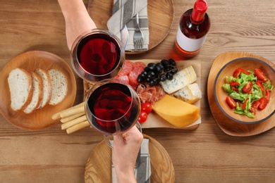 People holding glasses of red wine over table with delicious food, top view