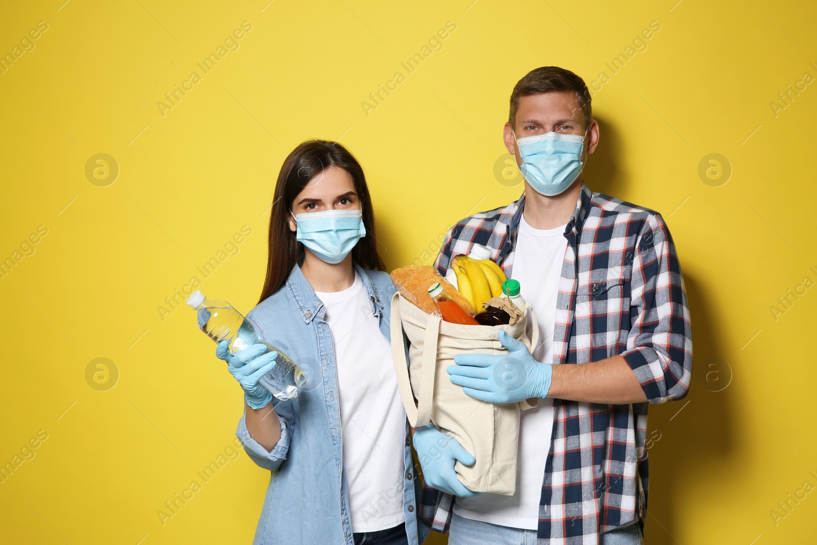 Photo of Volunteers in protective masks and gloves with products on yellow background. Aid during coronavirus quarantine