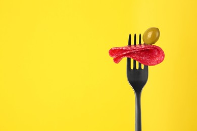 Photo of Fork with tasty slice of salami and olive on yellow background, space for text