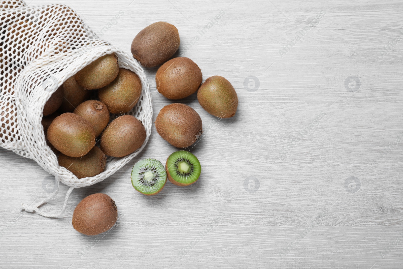 Photo of Net bag with cut and whole fresh kiwis on white wooden table, flat lay. Space for text