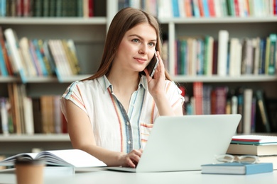 Photo of Young woman talking on phone and working with laptop at table in library