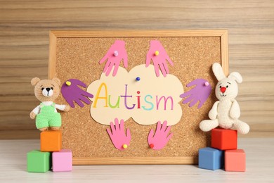 Cork board with word Autism, paper hands and different toys on white wooden table