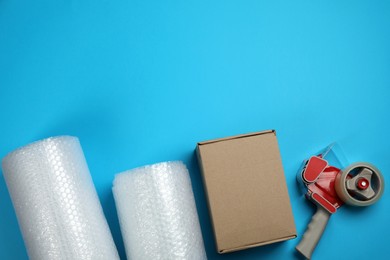 Photo of Bubble wrap rolls, cardboard box and tape dispenser on light blue background, flat lay. Space for text