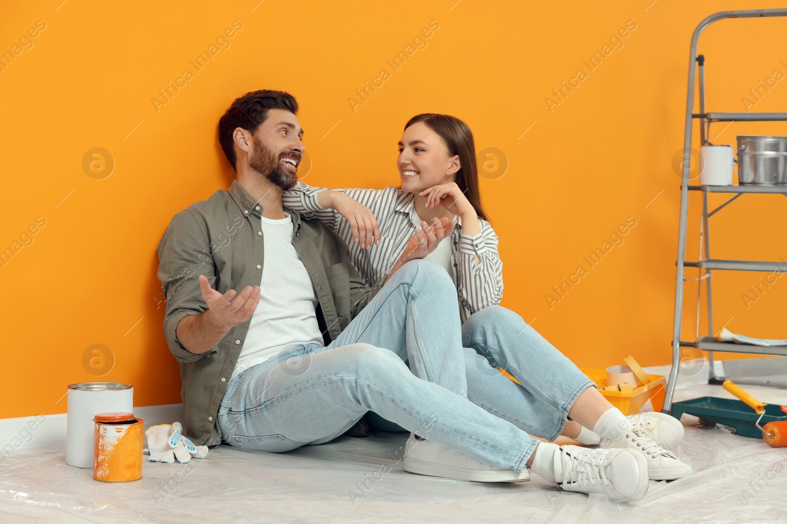 Photo of Happy designers sitting on floor with painting equipment near freshly painted orange wall indoors