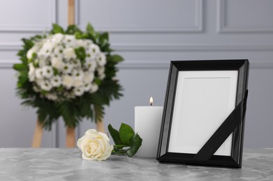 Photo of Photo frame with black ribbon, rose, burning candle on light grey table and wreath of flowers near wall indoors, space for text. Funeral attributes