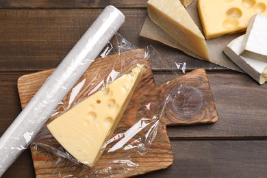 Different cheeses and plastic food wrap on wooden table, flat lay