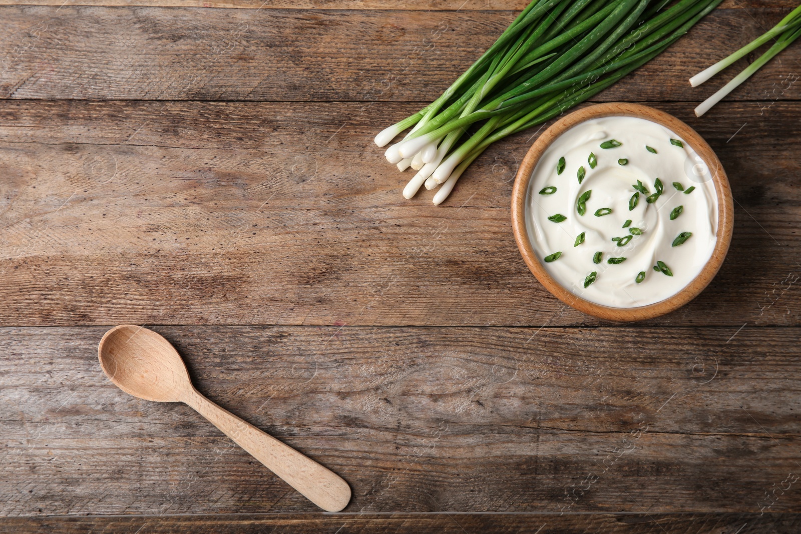 Photo of Bowl of fresh sour cream with green onion and spoon on wooden table, flat lay. Space for text