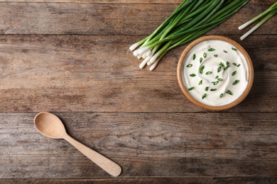 Photo of Bowl of fresh sour cream with green onion and spoon on wooden table, flat lay. Space for text