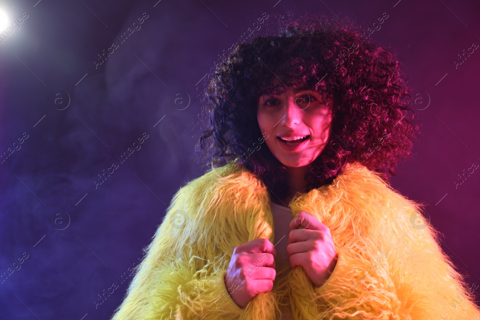 Photo of Beautiful young woman in yellow fur coat on color background in neon lights and smoke