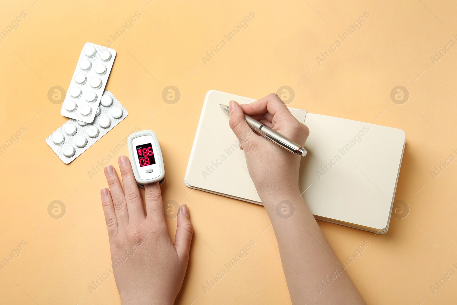 Photo of Woman with pen and notebook using fingertip pulse oximeter on light orange background, flat lay