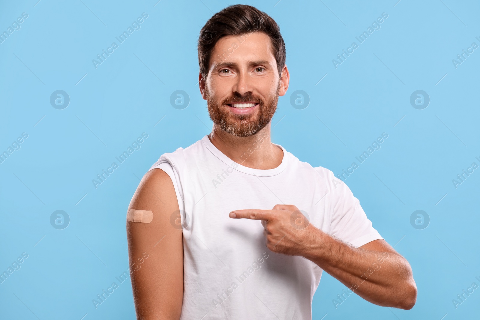 Photo of Man pointing at sticking plaster after vaccination on his arm against light blue background