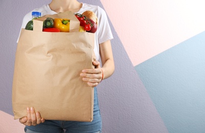 Photo of Woman holding paper bag with different groceries near color wall, closeup view. Space for text