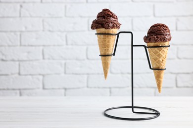 Chocolate ice cream scoops in wafer cones on white wooden table against brick wall, space for text