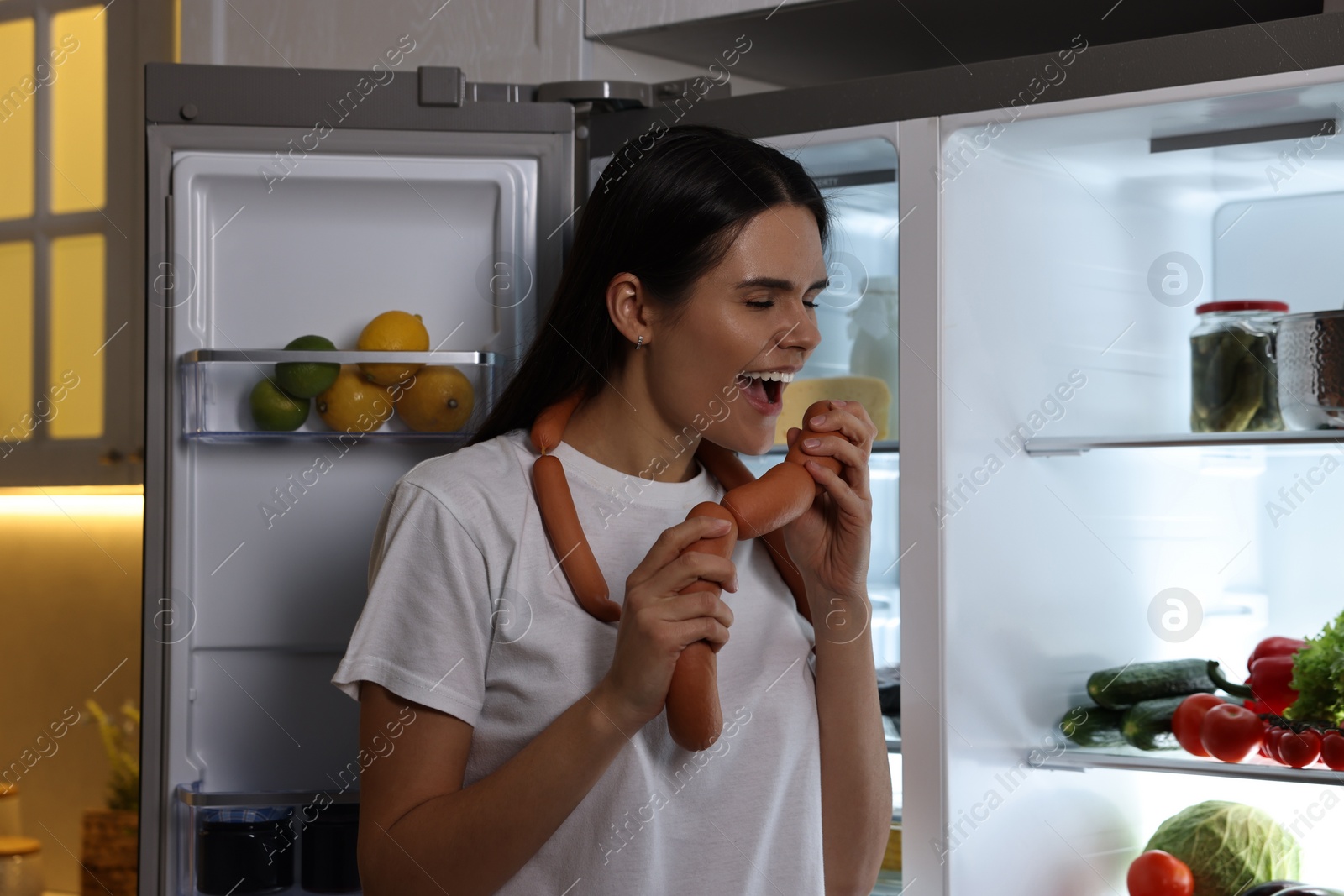 Photo of Young woman eating sausages near modern refrigerator in kitchen at night