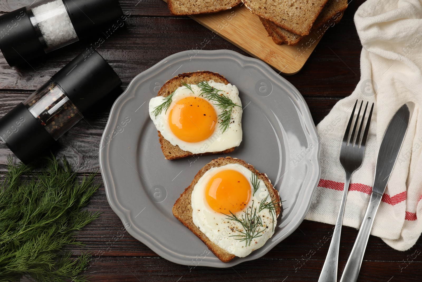Photo of Plate with tasty fried eggs, slices of bread, dill, fork and knife on dark wooden table, flat lay