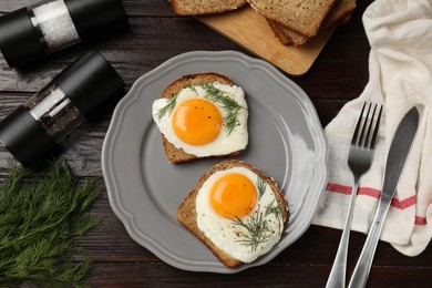 Plate with tasty fried eggs, slices of bread, dill, fork and knife on dark wooden table, flat lay