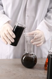 Photo of Woman pouring black crude oil from beaker into flask at grey table, closeup