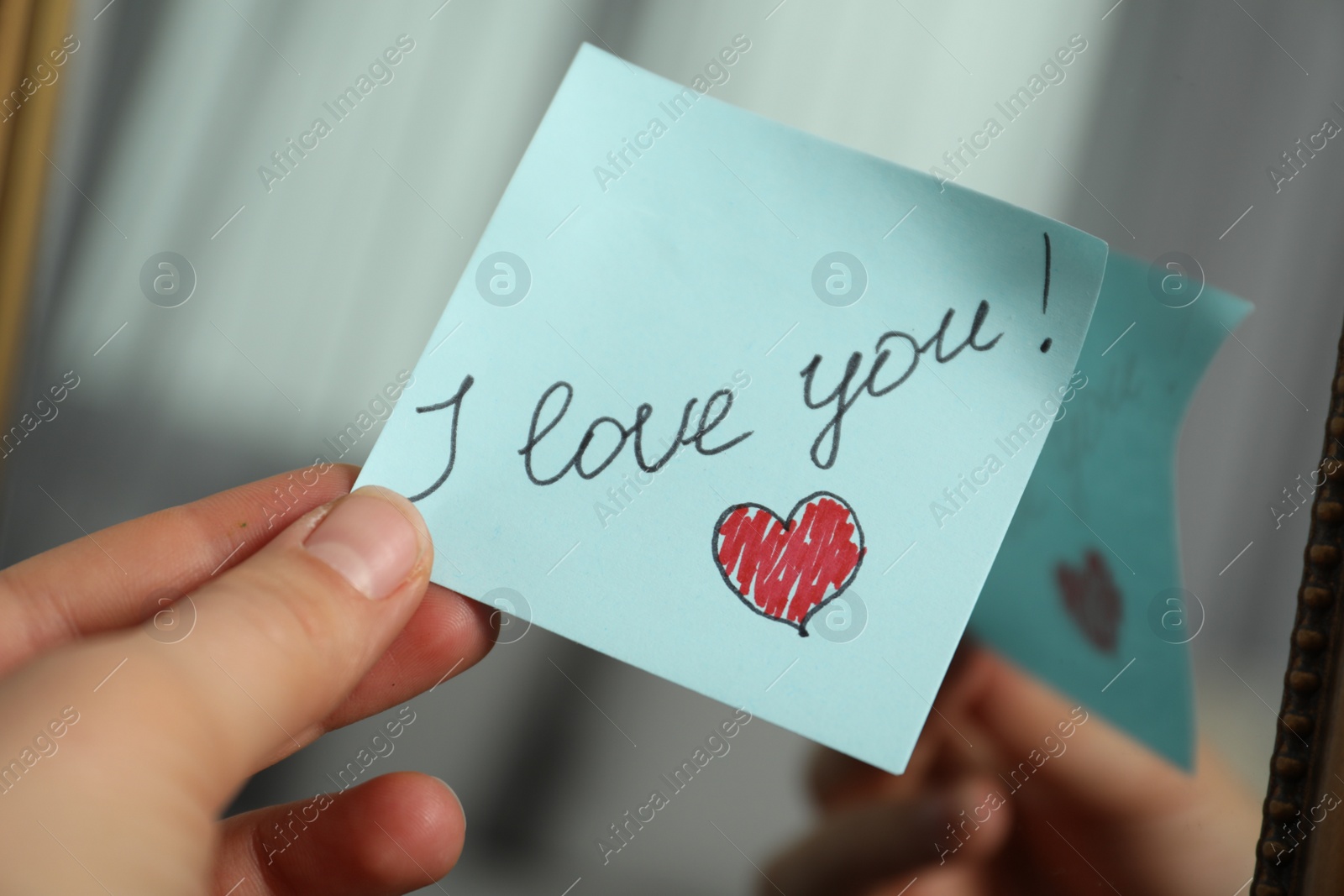 Photo of Woman holding sticky note with phrase I Love You near mirror, closeup