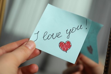 Photo of Woman holding sticky note with phrase I Love You near mirror, closeup