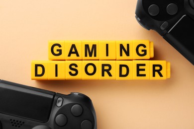 Phrase Gaming Disorder made of yellow cubes and gamepads on beige background, flat lay. Addictive behavior