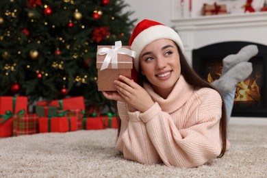 Happy young woman in Santa hat with gift box in room decorated for Christmas