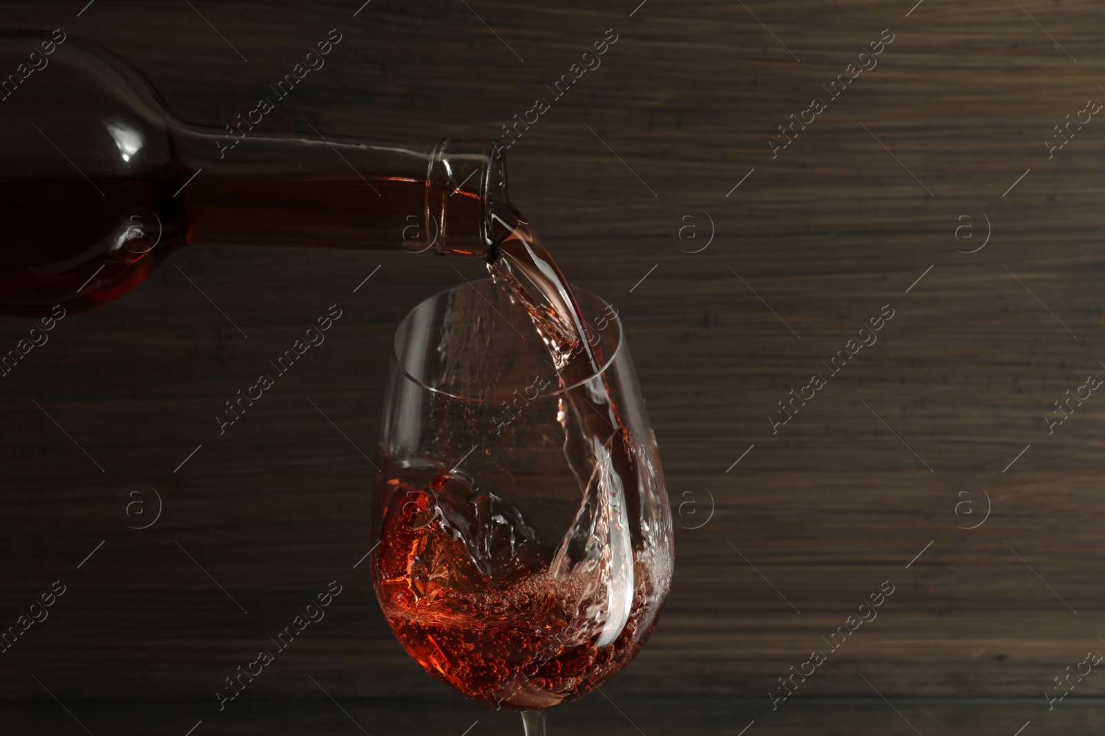 Photo of Pouring rose wine from bottle into glass against wooden background, space for text