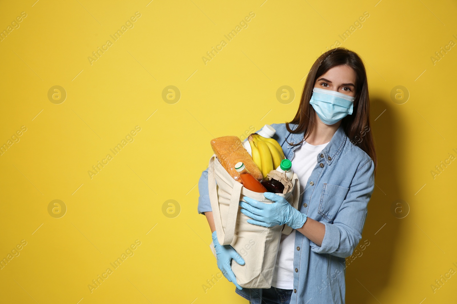Photo of Female volunteer in protective mask and gloves with products on yellow background, space for text. Aid during coronavirus quarantine