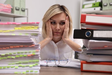 Overwhelmed woman sitting at table with stacks of documents and folders in office