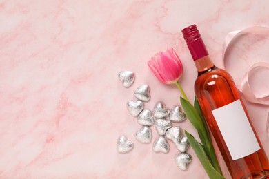 Delicious heart shaped chocolate candies, beautiful tulip and bottle of wine on pink table, flat lay. Space for text