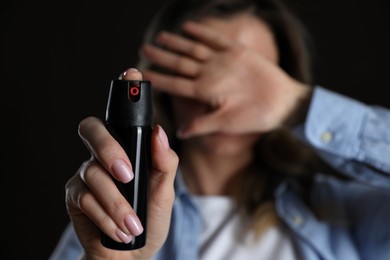 Young woman covering eyes with hand and using pepper spray on black background, focus on canister