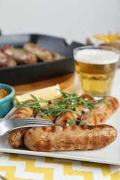 Photo of Tasty grilled sausages served with arugula and beer on table, closeup