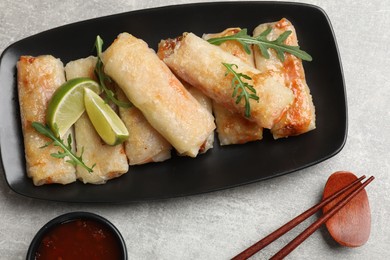 Photo of Tasty fried spring rolls, arugula, lime and sauce served on grey textured table, flat lay