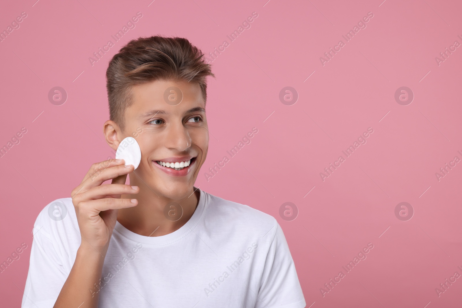 Photo of Handsome man cleaning face with cotton pad on pink background, space for text