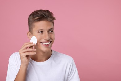 Handsome man cleaning face with cotton pad on pink background, space for text