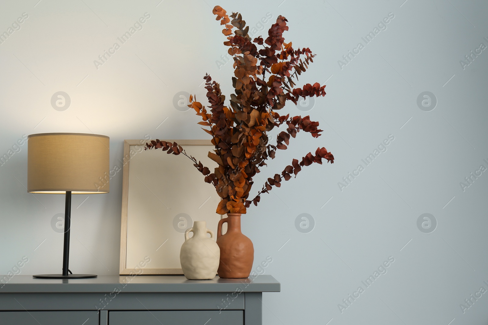 Photo of Stylish vases, dried eucalyptus branches and table lamp on chest of drawers near white wall indoors, space for text. Interior design