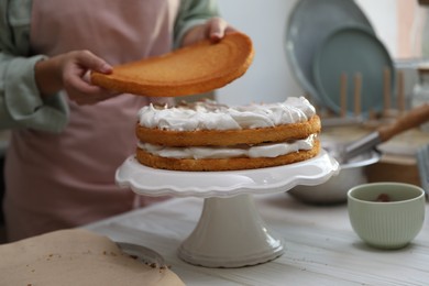 Photo of Woman stacking layers of sponge cake at white wooden table in kitchen, closeup