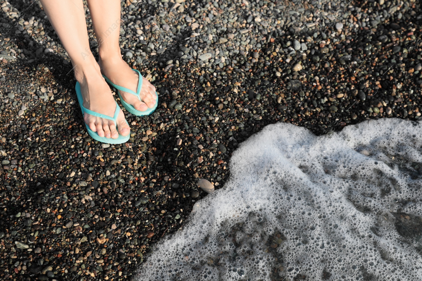 Photo of Woman in stylish flip flops on pebble beach near beautiful sea wave, closeup