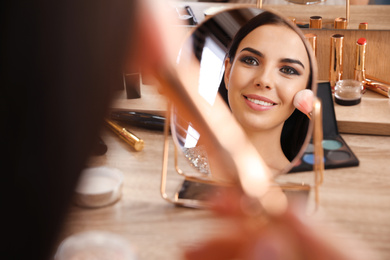 Photo of Beautiful young woman applying makeup near mirror at dressing table