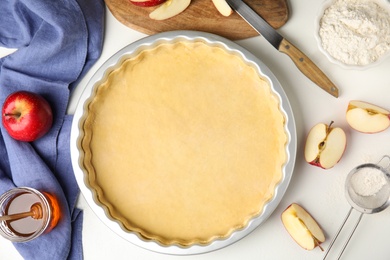 Photo of Baking dish with dough for traditional English apple pie and ingredients on white table, flat lay