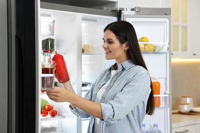 Photo of Young woman taking bottle of ketchup out of refrigerator in kitchen
