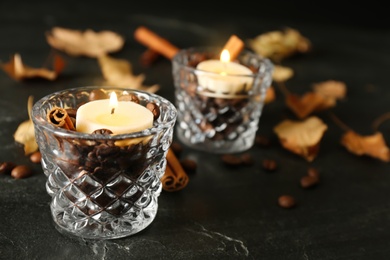 Glass holders with burning candles, coffee beans and cinnamon on black stone table, closeup. Space for text