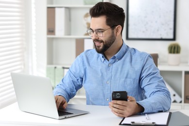 Photo of Handsome young man using smartphone while working with laptop at white table in office