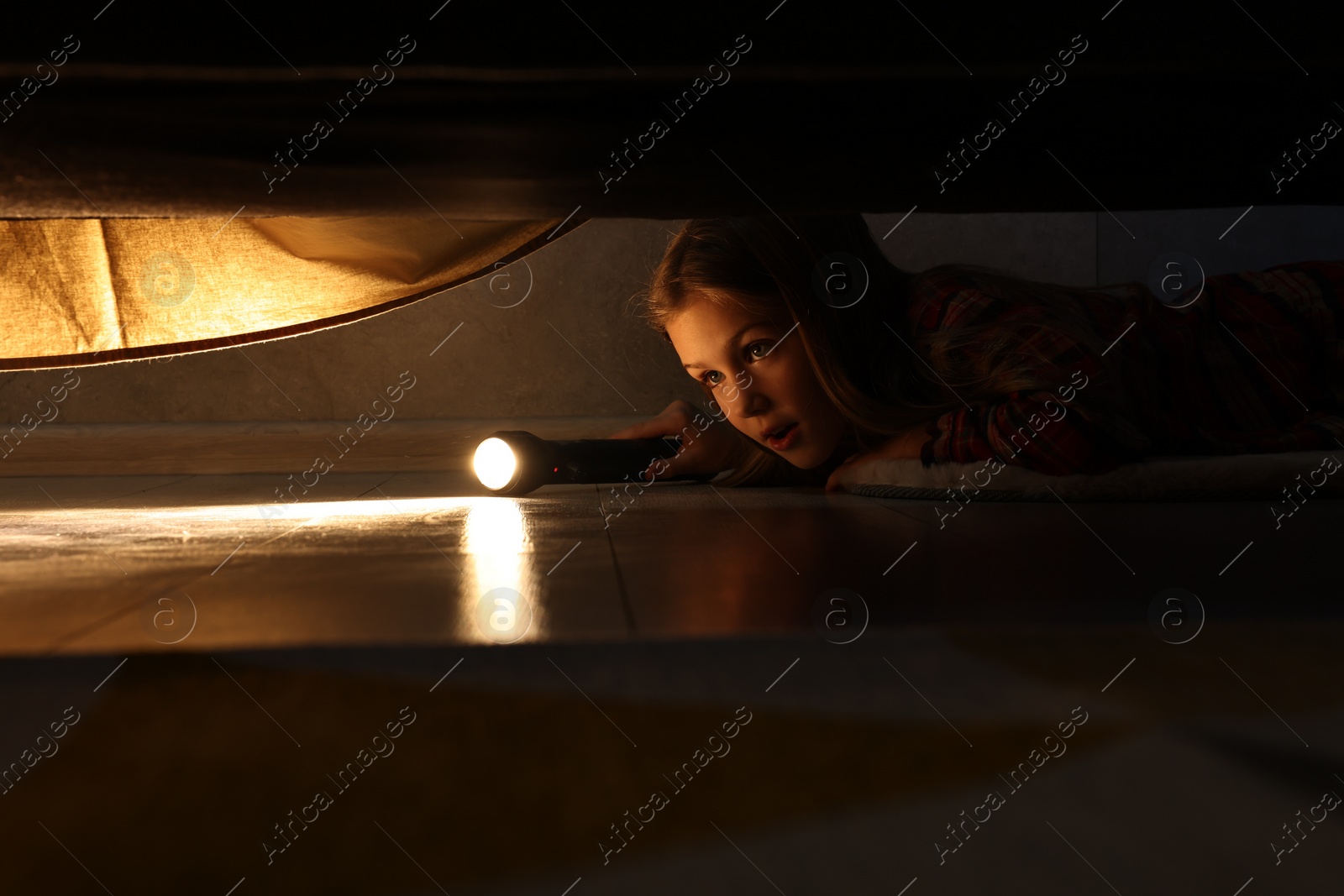 Photo of Little girl with flashlight looking for monster under bed at night