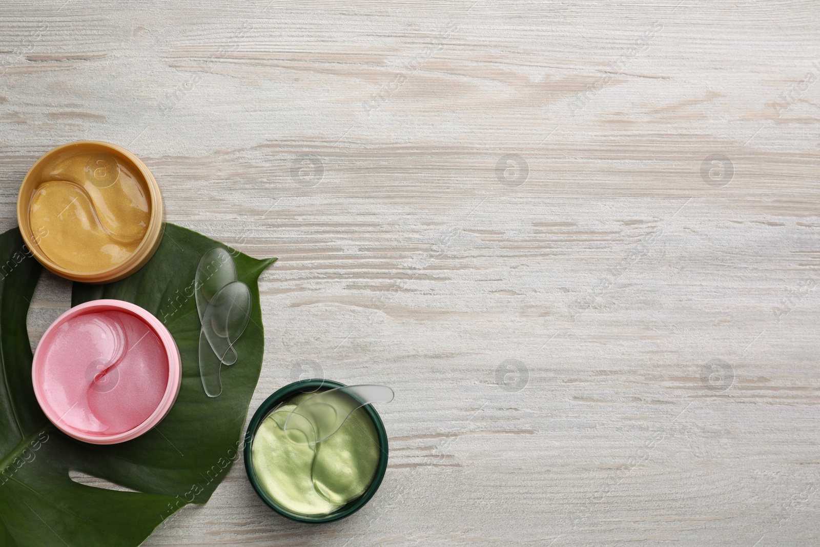 Photo of Packages of different under eye patches and tropical leaf on wooden table, flat lay with space for text. Cosmetic product