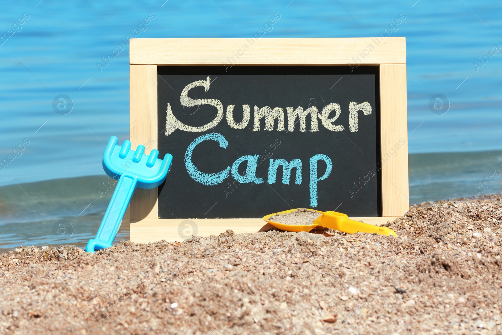 Photo of Little blackboard with text SUMMER CAMP and toys on sand near sea